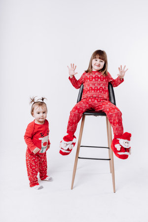 Merry Christmas and Happy Family Holidays. Cheerful cute children girls wearing in Xmas red pyjamas having fun on white background. Smiling older sister sitting on chair and playing. Loving sibling.の写真素材