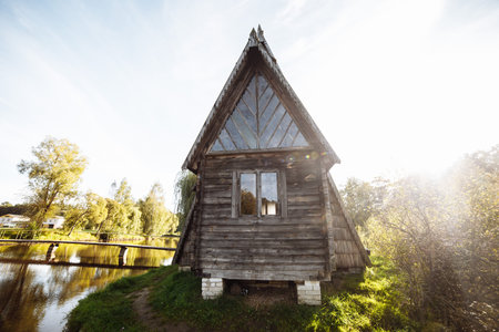 An old wooden village house stands on the edge of the forest by the lake against the blue sky. old village hut. Peasant dwelling. Abandoned housing people abandoned the houseの写真素材