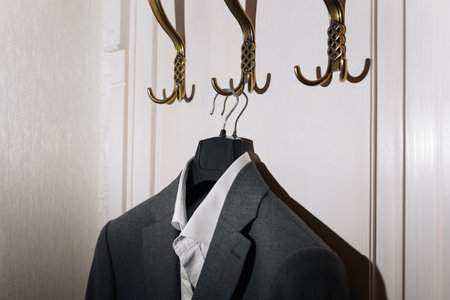 Man's dark gray suit jacket and white shirt hanging on gold metal hook on white wall backgroundの写真素材