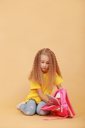 Children and modern technologies. Curly schoolchild girl is putting a digital tablet in her pink backpack, studying or playing game sitting on yellow orange studio background, free copy spaceの写真素材