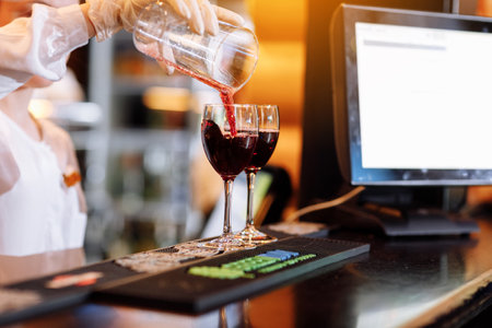 Bartender is pouring red wine into two long-stemmed wineglasses from a measuring cup on the bar counter. Blurred background. Party , cocktail, alcohol, beverage, restaurant, relax conceptの写真素材
