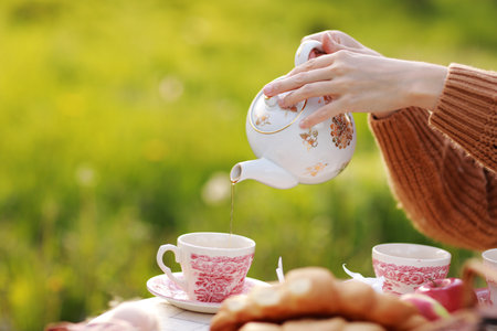 female hands pour tea into a cup from a teapot. Breakfast picnic with croissants and tea in spring blooming park on a white tablecloth on a sunny day. Outdoor, picnic, brunch, spring mood.の写真素材