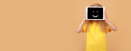 great application. Banner girl holding and showing digital tablet with screen with smile happy emoticon, covering face, recommending new app posing standing on yellow studio background. copyspace.の写真素材