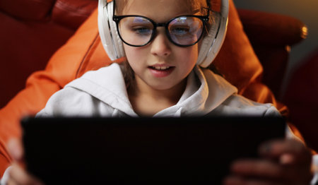 Excited teen girl plays game at home on digital tablet technology device sitting on sofa. Emotional child in headphones and glasses holds pad computer surfing internet.Children tech addiction concept.の写真素材