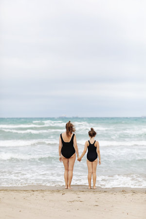 Happy family on beach vacation. Back view of mother and little daughter in sweatsuits are standing together and holding hands on beach. Mom and child girl kid enjoy and having fun at summer holidays.の写真素材