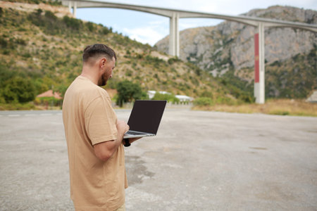 Collected man in casual clothes working on laptop outdoors on bridge construction background. Concept of remote work engineer or freelancer lifestyle. Cellular network broadband coverage. internet 5G.の写真素材