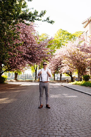 Stylish smiling young man wearing sunglasses, classic white shirt, gray pants and suspenders is standing at city street in the park with blooming pink cherry trees or sakura on spring warm day.の写真素材
