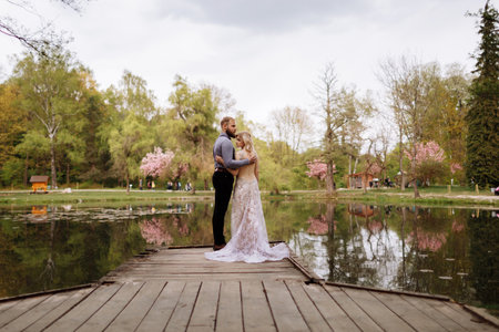 Side view of beautiful stylish newlyweds couple, bearded groom and bride in luxury long white dress are hugging by the pond in blooming pink cherry, sakura blossom park. Wedding day, valentines day.の写真素材