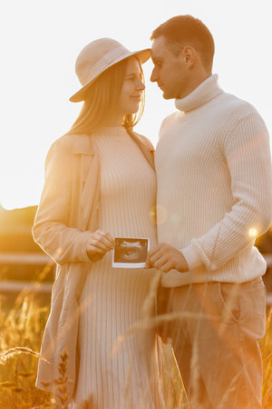 Unrecognized of a pregnant woman with her husband are holding an ultrasound scan photo of the unborn child outdoors on nature in sunlights. Happy motherhood and parenthood concept.の写真素材