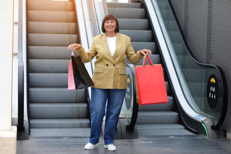 Stylish smiling adult woman is lifting down on escalator with colorful shopping bags. Attractive female spending leisure time at big modern mall. Online shopping concept, Seasonal Sales.Black Friday.の写真素材