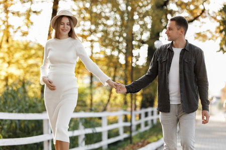 Pregnancy and Parenthoods concept - smiling man is walking with pregnant wife and looking to each other in autumn park. Happy styling young family is relaxing outdoors on weekend on sunny dayの写真素材