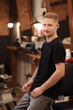 Stylish barber with beard and mustache man in casual work clothes holds professional scissors for haircut stands in hair salon. Confident hairdresser stands near workplace in barbershop. beauty salon.の写真素材