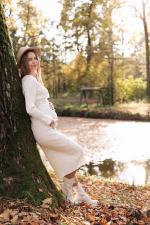 Elegant young pregnant woman in white knit dress and hat touching and stroking belly, relaxing in park by the lake on autumn warm day. Preparation for childbirth, healthy motherhood. Mothers day.の写真素材