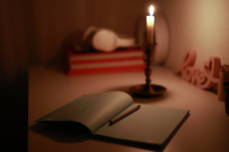 In the dark room, burning candle in candlestick is lighting on the work desk where the paper notebook with pen. Studying and working at home at blackout. Problem with electricity.の写真素材