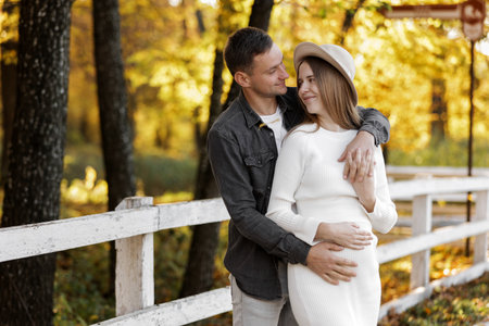 Happy stylish pregnant woman and man hugs, kiss, touches belly and listen baby's kicks, walk outdoor in autumn park. Pregnancy, motherhood and parenthood concept. Family is expecting for unborn babyの写真素材