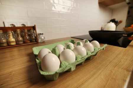 White chicken eggs in green cardboard egg tray on tabletop at modern stylish kitchen. Black frying wok pan on induction stove. Delicious healthy simple breakfast meal. Natural egg protein to the diet.の写真素材