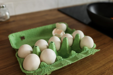 White chicken eggs in green cardboard egg tray on tabletop at modern stylish kitchen. Black frying...