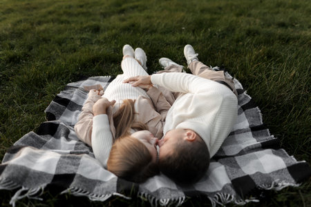 Happy family day. Top view of man and pregnant wife are embracing, lying and kissing on plaid on grass outdoors. mother's day. father's day. Pregnancy, parenthood, motherhood, love conceptの写真素材