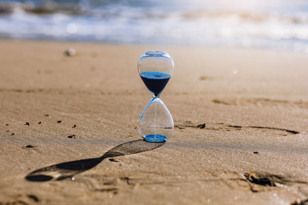 Hourglass with blue sand inside in a warm golden morning sunlight with shadow on a sandy beach and sea background, starting time for a new day or running of time. Concept of the rapid passage of time.の写真素材