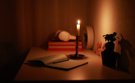 In the dark room, burning candle in candlestick is lighting on the work desk where the paper...