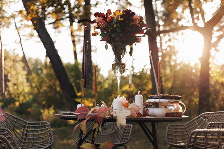Cozy autumn picnic in the park. Close up of table setting with white plates, cutlery, glass vase with colorful wildflowers, homemade apple pie, maple leaves, burning candles on wooden table outdoorsの写真素材
