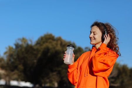 Active young woman in wireless headphones drinks water from bottle after exercising outdoors at...