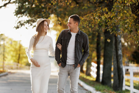 Pregnancy and Parenthoods concept - smiling man is walking with pregnant wife and looking to each other in autumn park. Happy styling young family is relaxing outdoors on weekend on sunny dayの写真素材