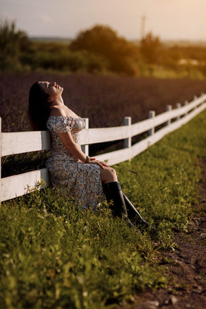 Young beautiful woman in floral sexy dress holds lavender flower in hands sits by the white wooden fence in blooming purple lavender field. Girl enjoying life and dreaming. natural beauty concept.の写真素材