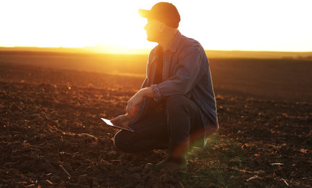 Agronomist touches soil examine and testing cultivated land before sowing on agricultural field and typing results in digital tablet on sunset. Smart farming technology and organic agricultureの写真素材