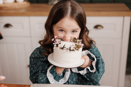 Adorable little child girl wears festive dress takes a bite out of a decorated flowers name cake at a birthday party.Happy smiling kid licks white cream from her dirty face and shows thumb up indoors.の写真素材