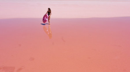 aerial view. Young woman in black swimsuit carries an inflatable pink float in her hands have fun dancing on the pink salt lake.Concept summer festivals, holiday, travel vacation, freedom, sun, enjoy.の写真素材