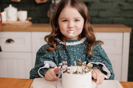 Adorable little child girl wears festive dress takes a bite out of a decorated flowers name cake at...