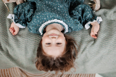 child birthday celebration. Top view of happy little girl 4-5 years wears stylish rustic style dress is lying on bed, smiling and looking at cameraの写真素材