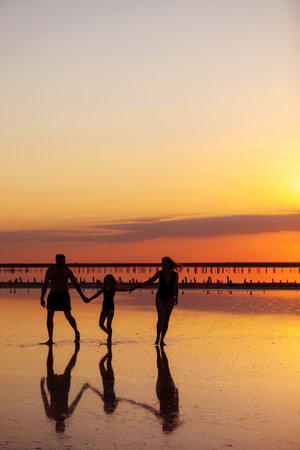 Happy family day. Silhouette of young mom, dad and little daughter holding hands walking together on beach on sunset in summer travel holiday. International Childrens Day. Concept of family valuesの写真素材