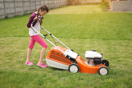 Cute teenage child girl working in garden, moving green grass with lawn-mower on backyard on summer holiday. Concept of pocket money, first salary. Special equipment for landscape, terrain. gardening.の写真素材