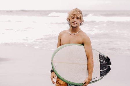 Handsome fit young blond man with mock up surfboard waits for wave to surf spot at sea ocean beach with black sand and looks at camera. Concept of sport, fitness, freedom, happiness, new modern life.の写真素材