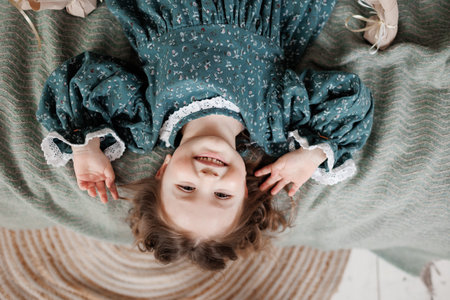 child birthday celebration. Top view of happy little girl 4-5 years wears stylish rustic style dress is lying on bed, smiling and looking at cameraの写真素材