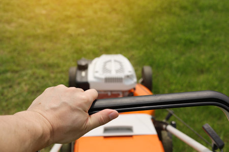 Cropped photo of professional male gardener is trimming and caring green grass lawn pushing modern electric cordless lawnmower at backyard of house. Landscaping industry themeの写真素材