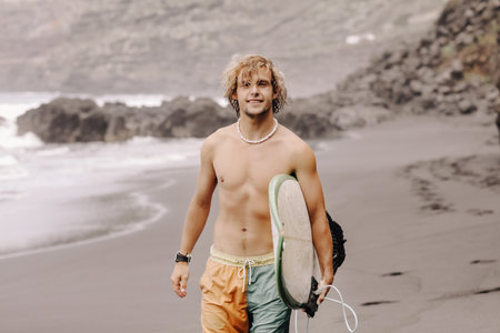 Handsome fit young blond man with mock up surfboard waits for wave to surf spot at sea ocean beach with black sand and looks at camera. Concept of sport, fitness, freedom, happiness, new modern life.の写真素材