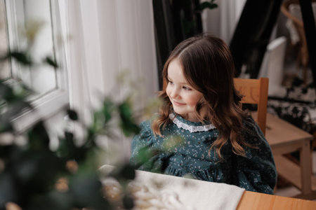 Adorable little girl with curly hair wears festive rural style dress is smiling, sitting by the table and supports her chin with hand at home. Happy childhood concept. Children day.の写真素材