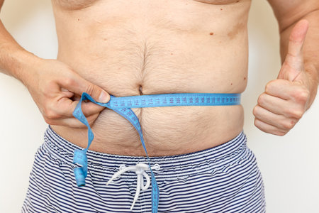 Man measures his fat belly with measuring tape and shows thumb up. Concept of weight loss, health problems of obese people. Controlling eating and active lifestyle. World Obesity Day.の写真素材