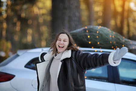 Young happy woman with rise up hands stands near gray car with Christmas tree in net and garlands on...