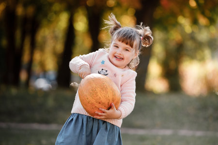 Cheerful little child girl lift very large orange pumpkin for her Halloween or Thanksgiving decoration outdoor at warm autumn day. good harvest. harvest time. farm vegetables. happy childhood concept.の写真素材