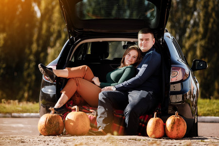 Couple in love, is sitting and hugging in car trunk at park with pumpkins decorated, on autumn warm day. Positivity and romantic man and woman celebrating thanksgiving holiday, Halloween. Fall time.の写真素材