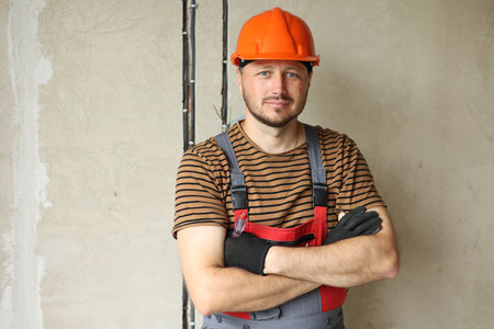 Renovation apartment Portrait of mature confident male foreman constructor repairman with crossed hands in safety hard hat and overalls looks at camera, holds screwdriver. Empty concrete wallsの写真素材