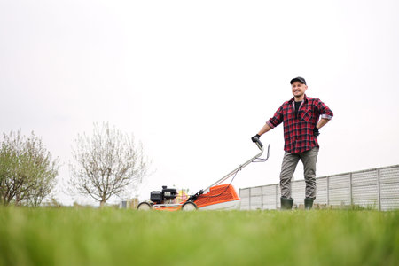 view of man in casual clothes mows lawn with lawn mower at the backyard of his house. Husband takes...