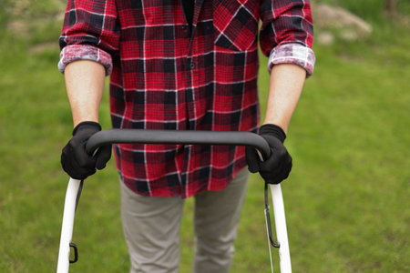Cropped photo of professional male gardener is trimming and caring green grass lawn pushing modern electric cordless lawnmower at backyard of house. Landscaping industry themeの写真素材
