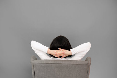 Rear view of unrecognizable woman with hands behind head sits on comfortable chair isolated on gray...