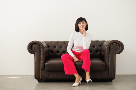Positive brunette middle aged woman wears elegant red pants, white blouse and high heels confidently looks at camera, sits relaxed on classic leather sofa at empty studio room. World women's day.の写真素材