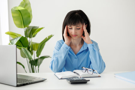 Overworked tired young businesswoman massages temples, suffers from headache after computer work sits at desk with laptop at office, exhausted female with closed eyes touching head, relieving painの写真素材